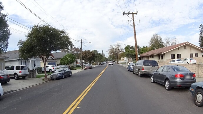 A two-lane street has a parking lane on either side. The parking lanes have no markings, and only the one going in this direction has any signs; the one going in the opposite direction is unrestricted. (© 2016 Bryan Housel, CC BY-SA 4.0.)