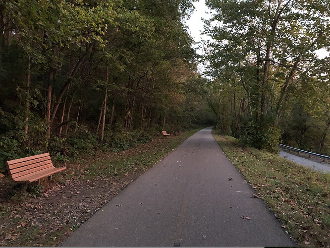 Two park benches about 80 feet (24 m) apart in a grassy area to one side of a paved off-road trail through a clearing in the woods. To the right of the trail is the Little Miami River, out of view just past the country road.