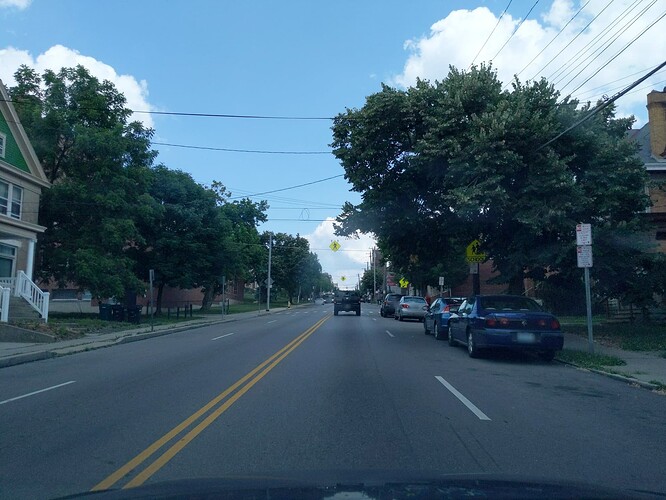 A four-lane street, two in either direction. Cars are parked all along the right lane. Regulatory signs say: No Stopping or Parking Except Loading, 10 Minute Limit, Tow Zone; No Stopping or Parking 7 AM to 6 PM, Monday through Saturday Except Truck Loading, Tow Zone. (© 2019 realadamp, CC BY-SA 4.0.)