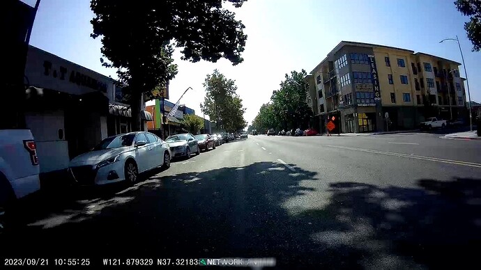 Another view of the same intersection shows a double yellow centerline that extends past a theoretical crossing of the main street from the curb ramp at the corner. A line of cars is parked at marked street parking stalls. (© 2023 networklanman, CC BY-SA 4.0.)